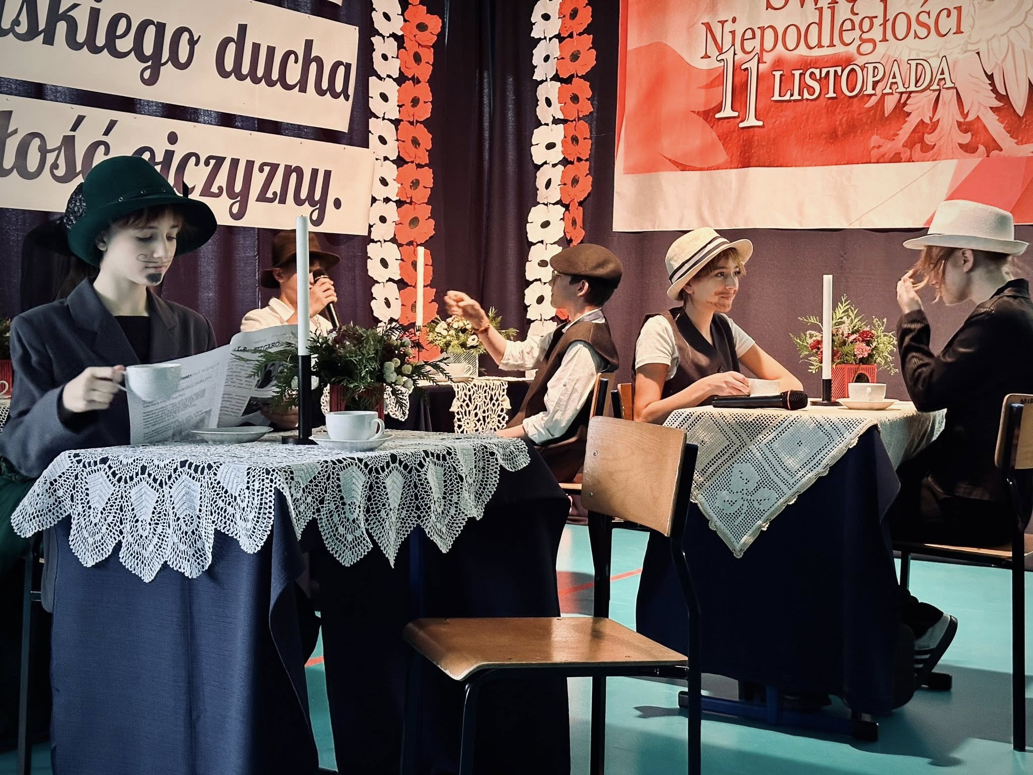 „Maria Skłodowska-Curie” czytająca gazetę w kawiarni. W tle widać innych gości przy stolikach.